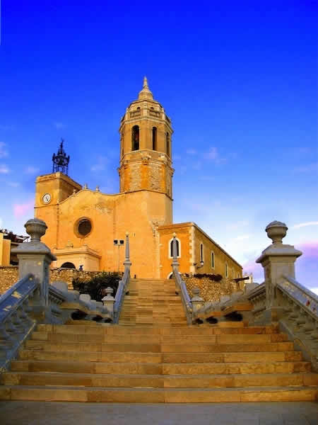 Iglesia de Sant Bartomeu y Santa Tecla en Sitges al atardecer, vista desde la escalinata con cielo azul claro.