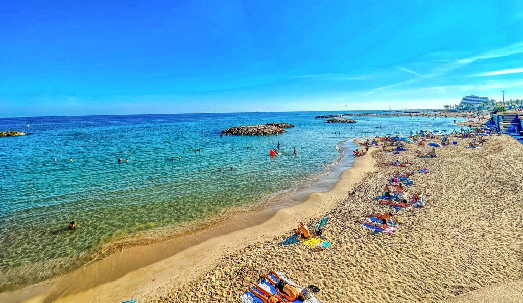 Playa de arena dorada en Sitges, con bañistas disfrutando del sol bajo un cielo despejado.